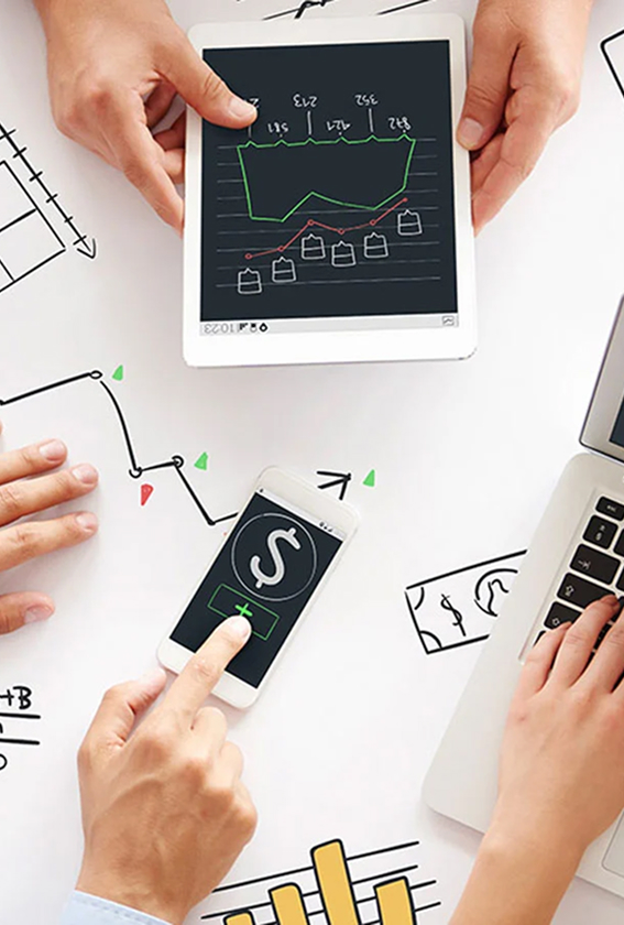 Top-down view of people collaborating on financial data using a tablet, smartphone with a dollar sign icon, and a laptop over a desk covered in hand-drawn charts